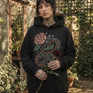 May include: A black hooded sweatshirt featuring a graphic print of a coiled snake and a large rose in muted tones of pink, green, and blue. The design includes small star accents. The hoodie has long sleeves, a front pocket, and a hood.