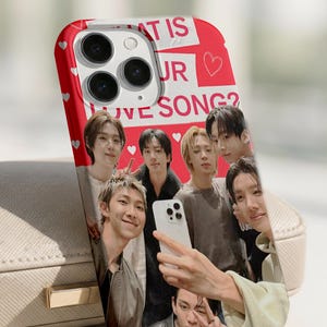 May include: A red phone case with a group of people posing for a selfie. The case has the text "WHAT IS UR LOVE SONG?" and small white hearts. The phone case is on a light-coloured surface.