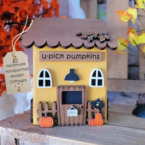 May include: A yellow wooden cottage decoration with a brown scalloped roof, featuring the text u-pick pumpkins, two white arched windows, a central door with an open sign, a small fence, orange pumpkin cutouts, and a black bird accent.