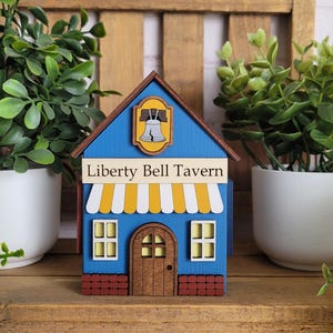 May include: A blue wooden house-shaped decoration with the text "Liberty Bell Tavern". The house has a yellow and white awning, a brown door, and a Liberty Bell emblem. It is flanked by potted plants.