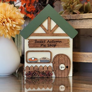 May include: A wooden decorative sign shaped like a small shop with a green roof, a brown door with an open sign, and a window displaying two pies. A wooden plaque reads Sweet Autumn Pie Shop, and a picket fence with autumn leaves sits below.