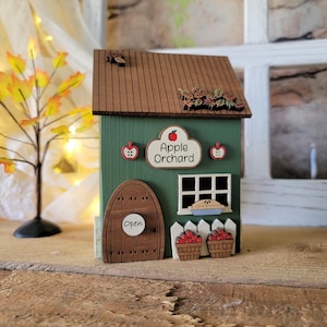 May include: A green wooden decorative house with a brown roof, featuring a sign that reads Apple Orchard, a rounded door with an Open label, a white window, two baskets of red apples, and a white picket fence with autumn leaf accents.