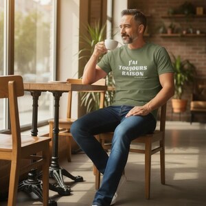 Könnte beinhalten: Ein Mann in einem grünen T-Shirt mit dem weißen Text "J'AI TOUJOURS RAISON", sitzt an einem Holztisch und hält eine weiße Kaffeetasse. Er trägt blaue Jeans und weiße Turnschuhe. Der Hintergrund ist ein Café mit einem großen Fenster.