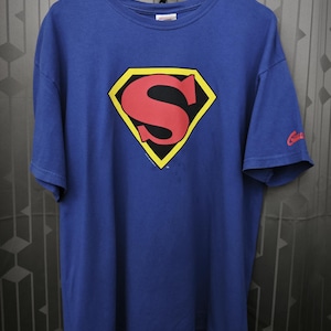 Könnte beinhalten: Ein blaues T-Shirt mit kurzen Ärmeln und dem ikonischen Superman-Logo auf der Brust, das aus einem roten S in einer gelb umrandeten schwarzen Rautenform besteht. Auf dem linken Ärmel befindet sich zudem ein kleines rotes Grafikdetail.