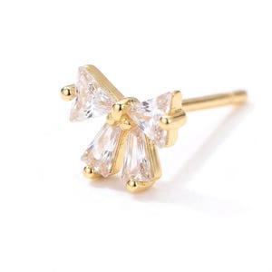 May include: A gold-coloured stud earring with a cluster of clear, sparkling cubic zirconia stones. The earring has a bow-like design with a post for pierced ears. The stones are cut in various shapes.