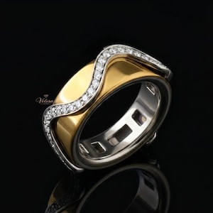 May include: A two-tone ring with a gold center band and a silver outer band. The gold band is accented with a wave-like design encrusted with small, clear gemstones. The ring is set against a black background.