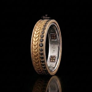 May include: A two-tone ring featuring a gold band with a textured chevron pattern alongside a row of small black stones. The interior of the silver-colored band displays the text Velora, 78, and TM, with two square-shaped decorative inlays.