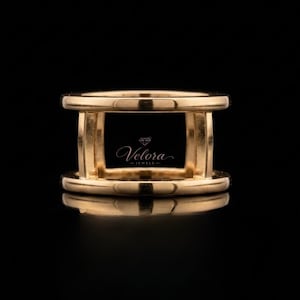 May include: A gold-colored ring with a rectangular design. The ring has a top and bottom band connected by vertical bars. The ring is highly reflective, with the brand name "Velora Jewels" visible in the center.