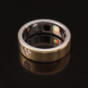May include: A gold-toned smart ring with a brushed finish, featuring a heart-shaped cluster of small clear stones on the outer band. The interior band is silver-toned and displays the Velora logo, a dark sensor square, and a glowing green light.