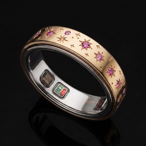 May include: A gold-toned smart ring featuring a celestial pattern of engraved starbursts and inlaid pink gemstones on the outer band. The interior surface is silver-toned and contains visible electronic sensors and the brand name Velora.