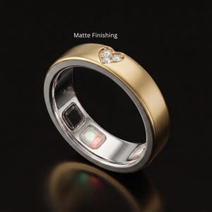 Puede incluir: Un anillo dorado con acabado mate y un detalle en forma de corazón que contiene tres pequeñas piedras claras. El interior de la banda presenta un sensor rectangular oscuro y un sensor cuadrado con luces verde y roja, además del texto Velora.