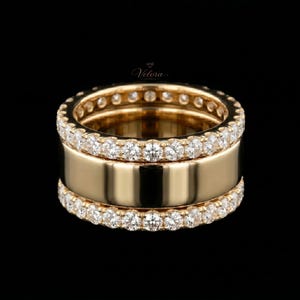 Könnte beinhalten: Ein goldener Ring mit einem breiten Band und zwei Reihen kleiner, klarer Edelsteine. Der Ring hat eine polierte, reflektierende Oberfläche und ist vor schwarzem Hintergrund platziert. Der Ring ist ein Schmuckstück.