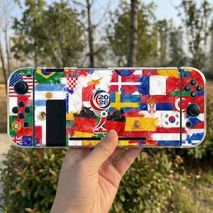 May include: A handheld gaming console featuring a protective skin decorated with a collage of various international flags and a central graphic logo displaying the text 2026. The device includes black control sticks, buttons, and a clear outer shell.