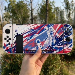 May include: A handheld gaming console featuring a custom skin with a dynamic soccer-themed graphic. The design displays a player in motion, a soccer ball, and a net, rendered in a vibrant color palette of red, white, and blue with abstract patterns.