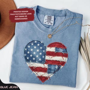 May include: A blue crew neck t-shirt featuring a printed heart graphic with a faux patchwork American flag design. The shirt has a Comfort Colors label and a thin gold-tone necklace with a small circular pendant resting on the neckline.