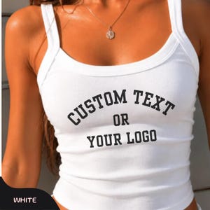 May include: A white ribbed tank top with thick shoulder straps and a scoop neckline. The front of the garment features black printed text that reads CUSTOM TEXT OR YOUR LOGO in a bold, arched font style across the chest area of the shirt.