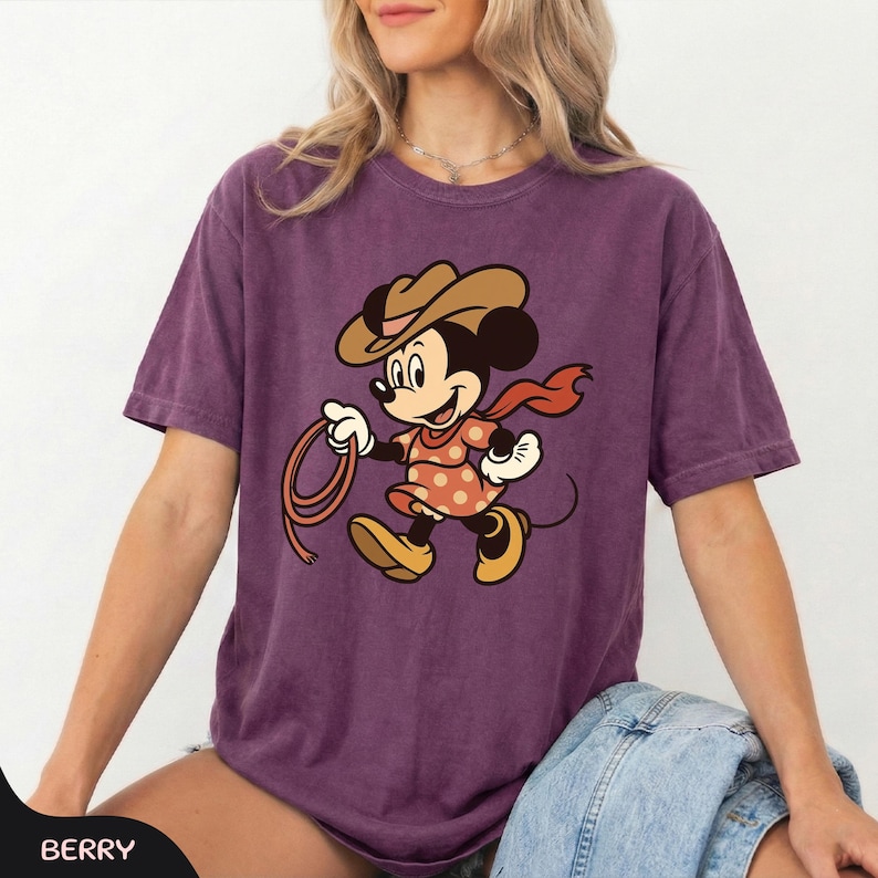 May include: Berry-colored t-shirt featuring a cartoon Minnie Mouse dressed as a cowgirl, wearing a brown hat, orange dress with white polka dots, and a red scarf. Minnie is holding a lasso. The shirt is a comfortable, casual style.