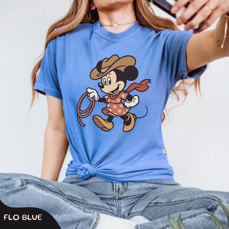 May include: A blue t-shirt featuring a cartoon character dressed as a cowgirl, wearing a brown hat, red scarf, and a polka-dot dress, holding a lasso. The text "FLO BLUE" is at the bottom of the image.
