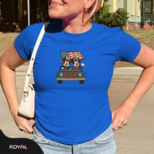 May include: Royal blue t-shirt featuring a graphic of Mickey and Minnie Mouse in a car with an American flag. The word "USA" is displayed below the car in a star-bordered font. The shirt is a crew neck.