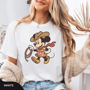 May include: White t-shirt featuring a cartoon Mickey Mouse dressed as a cowboy. The character wears a brown hat, a red scarf, and a polka-dot shirt, holding a lasso. The word "WHITE" is printed at the bottom of the image.
