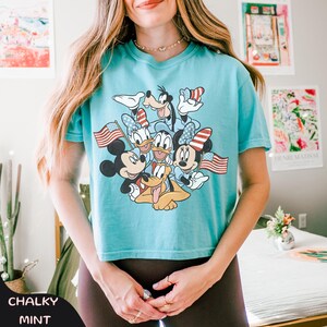 May include: A light blue t-shirt featuring a graphic of Mickey Mouse, Minnie Mouse, Donald Duck, Daisy Duck, Goofy, and Pluto, each holding an American flag. The shirt has the words "Chalky Mint" at the bottom.