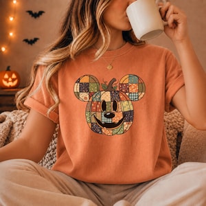 May include: An orange short-sleeve t-shirt featuring a graphic of a pumpkin shaped like a mouse head with a patchwork pattern. The pumpkin has a stem on top, a black nose, and a carved jack-o'-lantern smile. The shirt has a crew neckline.