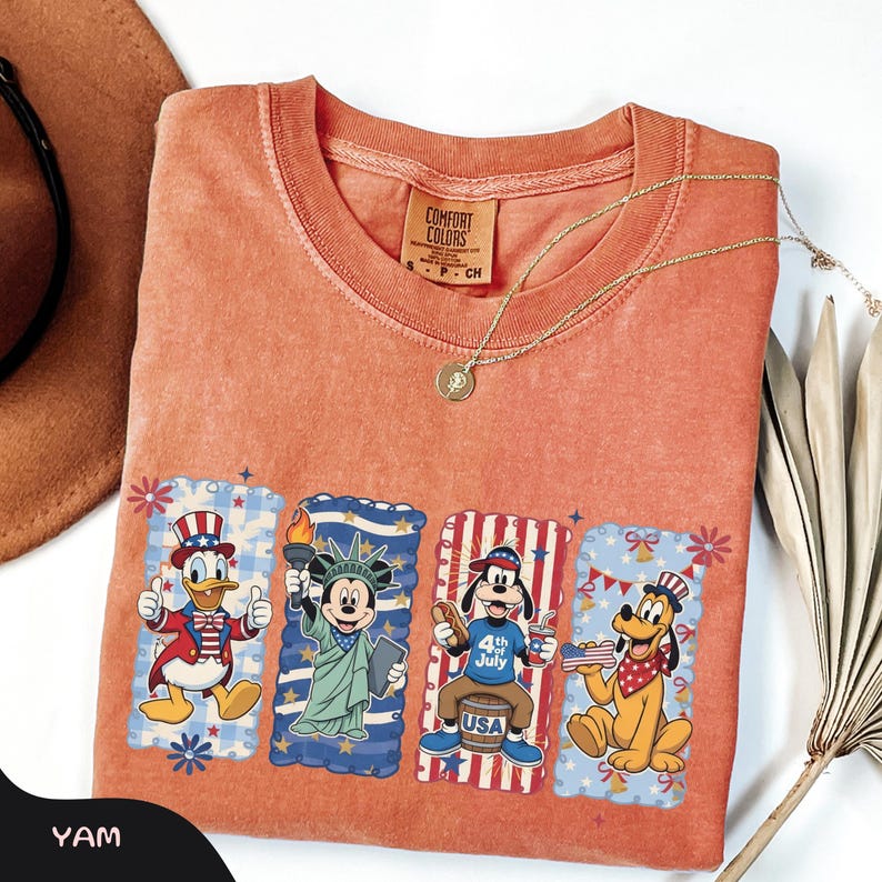 May include: An orange Comfort Colors t-shirt featuring four Disney characters in patriotic attire. Donald Duck, Mickey Mouse, Goofy, and Pluto are depicted with American flags and 4th of July themes. A gold necklace and a brown hat are also visible.