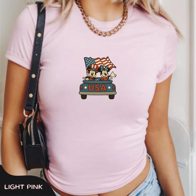 May include: Light pink t-shirt featuring a patriotic graphic of cartoon characters in a car with American flags. The word "USA" is displayed on the front of the car. The shirt has a crew neck and short sleeves.