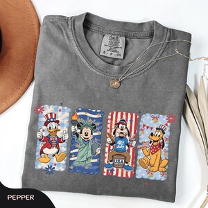 May include: A gray Comfort Colors t-shirt featuring four cartoon character designs. Donald Duck, Mickey Mouse, Goofy, and Pluto are depicted in patriotic scenes. A silver necklace with a pendant is draped across the shirt. The word "PEPPER" is at the bottom.