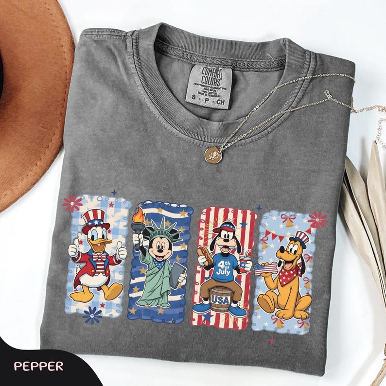 May include: A gray Comfort Colors t-shirt featuring four cartoon character designs. Donald Duck, Mickey Mouse, Goofy, and Pluto are depicted in patriotic scenes. A silver necklace with a pendant is draped across the shirt. The word "PEPPER" is at the bottom.