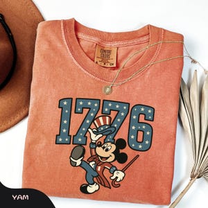 May include: Coral-colored t-shirt featuring the number "1776" in blue with white stars, and a cartoon character in patriotic attire. The shirt has a Comfort Colors tag and is accessorized with a gold necklace. A brown hat and dried plant are in the background.