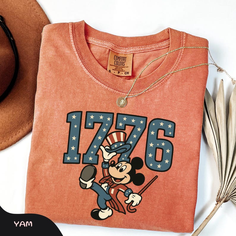 May include: Coral-colored t-shirt featuring the number "1776" in blue with white stars, and a cartoon character in patriotic attire. The shirt has a Comfort Colors tag and is accessorized with a gold necklace. A brown hat and dried plant are in the background.