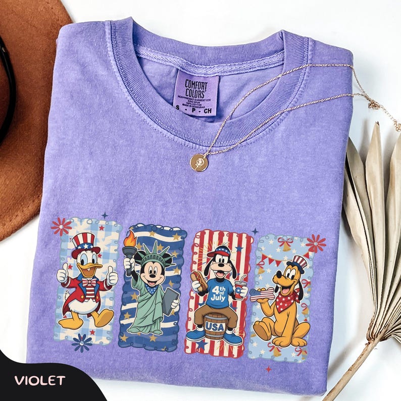 May include: Violet Comfort Colors t-shirt featuring patriotic Disney characters: Donald Duck, Mickey Mouse, Goofy, and Pluto. Each character is in a rectangular frame with red, white, and blue accents. A gold necklace and a brown hat are also visible.