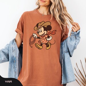 May include: Rust-colored t-shirt featuring a cartoon Minnie Mouse dressed as a cowgirl, complete with a brown hat, red scarf, and a lasso. The graphic is centered on the shirt, which has rolled-up sleeves. The word "YAM" is printed on the bottom left.