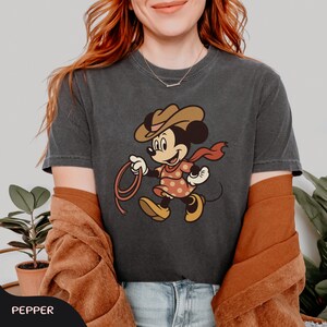 May include: Dark gray t-shirt featuring a cartoon Minnie Mouse dressed as a cowgirl, wearing a brown hat, red scarf, and a polka-dot dress. She is holding a lasso. The word "PEPPER" is at the bottom.