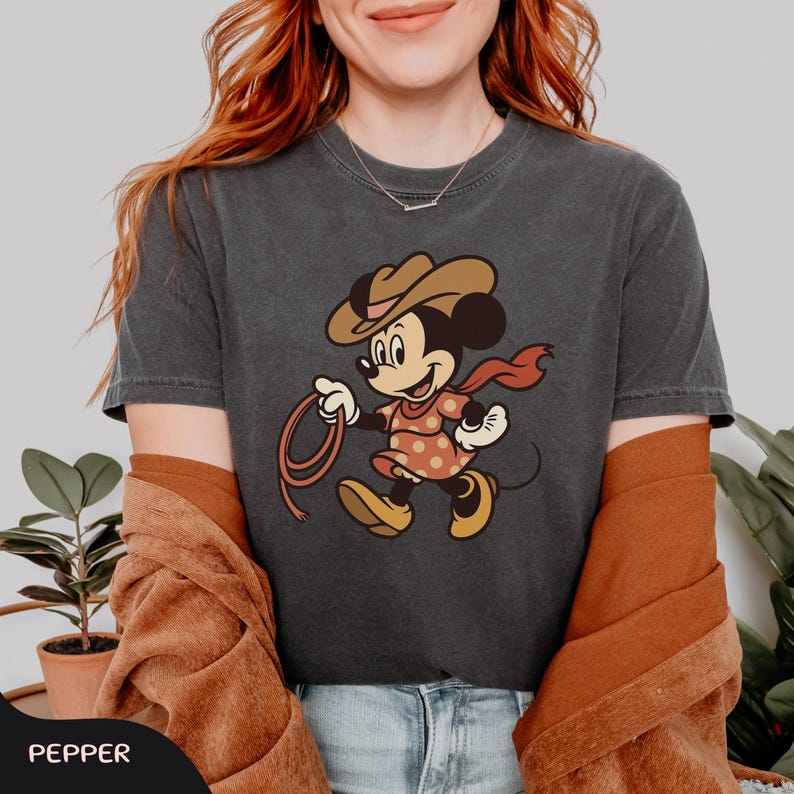 May include: Dark gray t-shirt featuring a cartoon Minnie Mouse dressed as a cowgirl, wearing a brown hat, red scarf, and a polka-dot dress. She is holding a lasso. The word "PEPPER" is at the bottom.