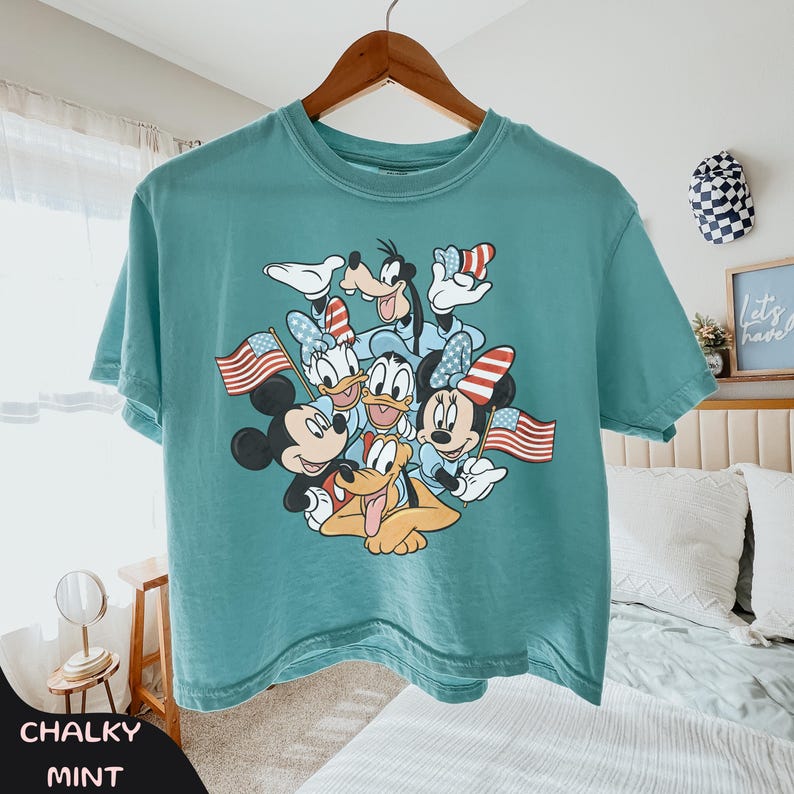 May include: A teal-colored t-shirt featuring a graphic of Disney characters, including Mickey and Minnie Mouse, Donald and Daisy Duck, Goofy, and Pluto. The characters are holding American flags and wearing patriotic accessories. The shirt is hanging on a wooden hanger.