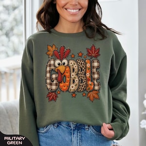 May include: A military green crewneck sweatshirt featuring a graphic of the word GOBBLE. The letters are filled with various patterns including plaid, leopard print, and pumpkins, with a turkey head integrated into the letter O.