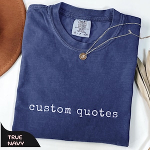 May include: A navy blue short-sleeved t-shirt with a crew neckline and the text "custom quotes" embroidered in white thread. The shirt features a Comfort Colors brand label at the inner neck and is paired with a thin gold-tone chain necklace.