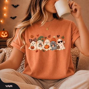 Könnte beinhalten: Ein orangefarbenes T-Shirt mit einer Grafik von fünf Cartoon-Geistern. Die Geister sind mit herbstlichen Elementen verziert, darunter eine karierte Schleife, eine grüne Mütze, ein Kürbis, ein karierter Hut und herzförmige Sonnenbrillen, umgeben von Blättern.