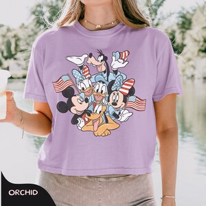 May include: A light purple t-shirt featuring a graphic of Mickey Mouse, Minnie Mouse, Donald Duck, Daisy Duck, Goofy, and Pluto. The characters are wearing patriotic accessories and holding American flags. The word "ORCHID" is printed at the bottom.