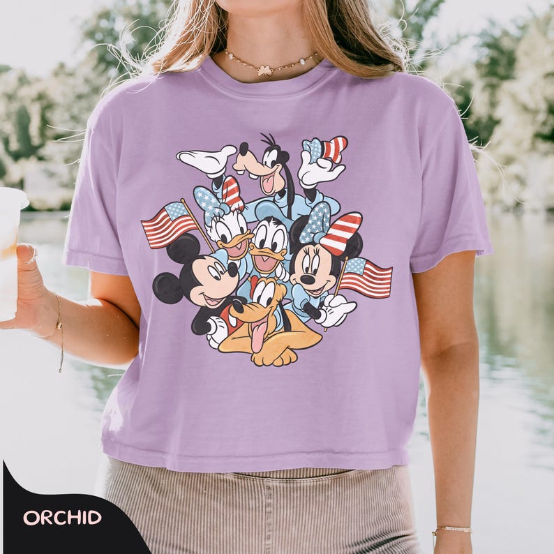 May include: A light purple t-shirt featuring a graphic of Mickey Mouse, Minnie Mouse, Donald Duck, Daisy Duck, Goofy, and Pluto. The characters are wearing patriotic accessories and holding American flags. The word "ORCHID" is printed at the bottom.