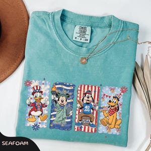 May include: Seafoam green t-shirt featuring four cartoon character designs celebrating the 4th of July. Donald Duck, Mickey Mouse, Goofy, and Pluto are depicted in patriotic attire. The shirt has a Comfort Colors label.