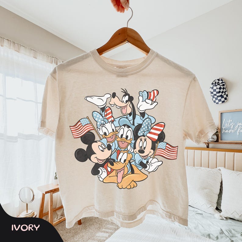 May include: Ivory-colored t-shirt featuring a graphic of Disney characters, including Mickey and Minnie Mouse, Goofy, Donald and Daisy Duck, and Pluto, with American flags and patriotic accents. The shirt is hanging on a wooden hanger.