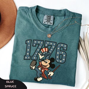 May include: Blue spruce t-shirt featuring the number "1776" with star accents and a cartoon character in patriotic attire. The shirt has a crew neck and a small tag. A gold necklace with a pendant is also visible.