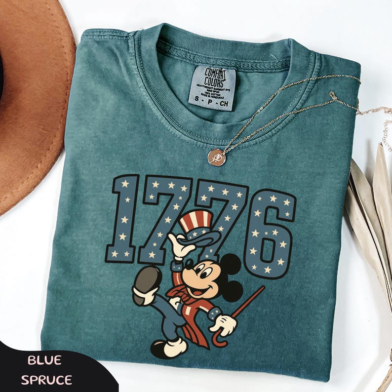 May include: Blue spruce t-shirt featuring the number "1776" with star accents and a cartoon character in patriotic attire. The shirt has a crew neck and a small tag. A gold necklace with a pendant is also visible.