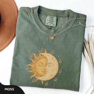 May include: A moss green t-shirt with a sun and moon graphic. The design features a golden sun and a cream-coloured moon embracing. A delicate gold necklace with a small pendant rests on the shirt. The shirt is labelled "Comfort Colors".
