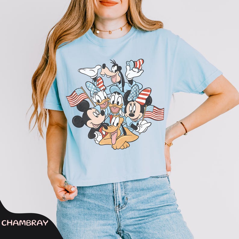 May include: Light blue t-shirt featuring a graphic of Mickey Mouse, Minnie Mouse, Donald Duck, Daisy Duck, Goofy, and Pluto holding American flags. The characters are wearing patriotic accessories. The shirt is made of a soft material.