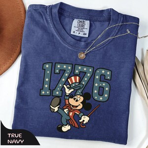 May include: A navy blue t-shirt with the text "1776" in a star-spangled design, featuring a cartoon character in patriotic attire. The shirt has a small tag that says "Comfort Colors". A gold necklace is draped across the shirt.