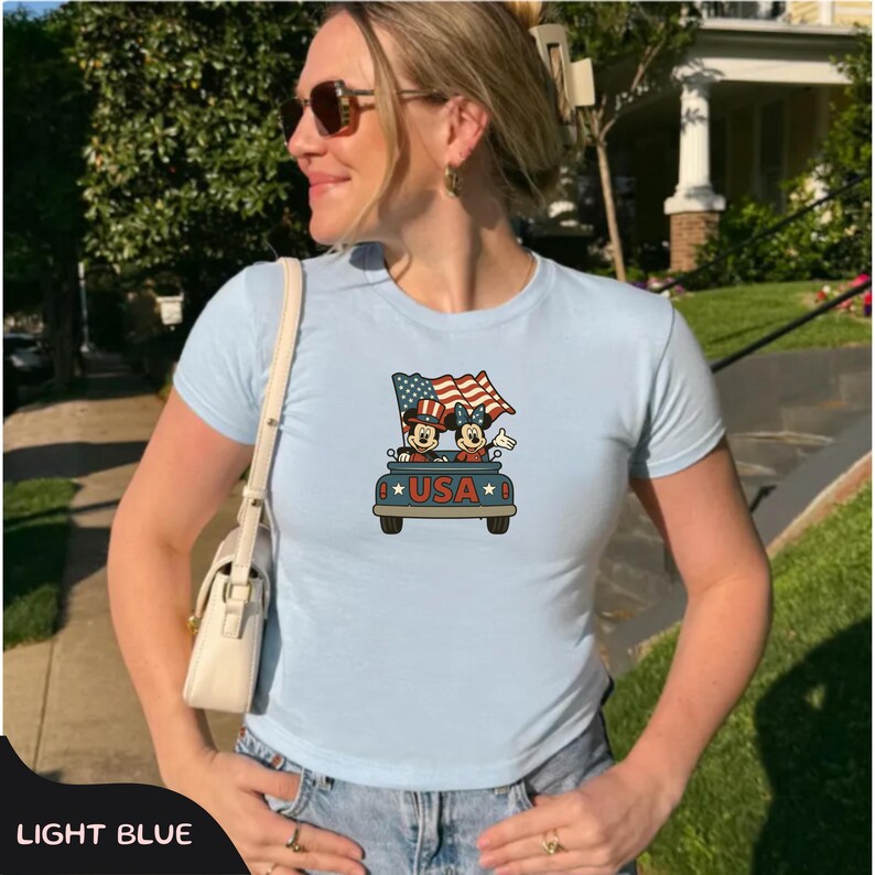 May include: Light blue t-shirt featuring a graphic of Mickey and Minnie Mouse in a car with American flags and the word "USA". The shirt has short sleeves and a crew neck.
